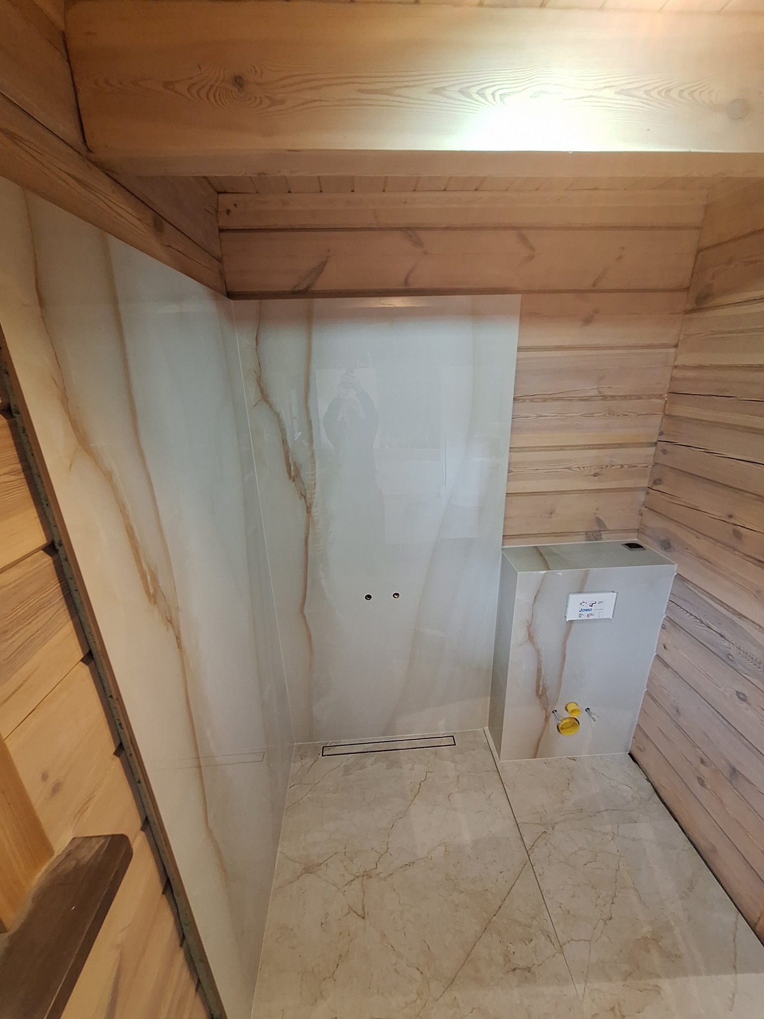 Łazienka wykończona drewnem i płytkami imitującymi marmur, widoczny podtynkowy stelaż WC i odpływ liniowy w podłodze.