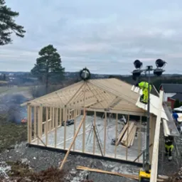 Wznoszenie drewnianej konstrukcji szkieletowej domu z wieńcem na dachu, widok z góry na plac budowy z elementami rusztowania i sprzętu budowlanego, w tle krajobraz z dymiącym ogniskiem.