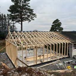 Stanislaw Domanski - Konstrukcja szkieletowa budynku w trakcie budowy, widoczne drewniane ramy ścian i dachu na betonowej płycie fundamentowej, teren wokół częściowo pokryty śniegiem, pochmurne niebo w tle.