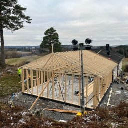 Stanislaw Domanski - Budowa domu szkieletowego w trakcie: widoczna drewniana konstrukcja, zarys ścian i dachu, teren budowy z elementami krajobrazu i sprzętem budowlanym, pochmurne niebo w tle.