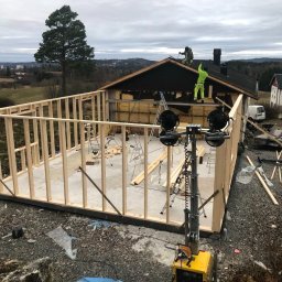 Stanislaw Domanski - Wznoszenie szkieletu domu jednorodzinnego, widoczna drewniana konstrukcja ścian, ocieplenie dachu, pracownicy na dachu, teren budowy z narzędziami i oświetleniem.