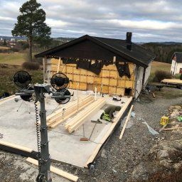 Stanislaw Domanski - Budowa domu: Widok z góry na plac budowy z fundamentami, izolacją ścian i stosem drewna, z pracownikiem w zielonym kombinezonie przycinającym drewno, w tle krajobraz z drzewami i zabudowaniami.