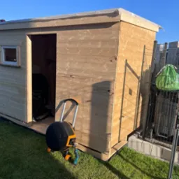 Surowa, drewniana konstrukcja małego domku ogrodowego z oknem, z widoczną drabiną opartą o bok i zwiniętym wężem ogrodowym przed wejściem, otoczona trawnikiem.