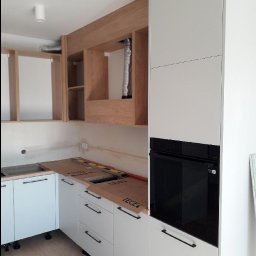 Stanislaw Domanski - Montaż kuchni na wymiar: białe szafki z czarnymi uchwytami, zabudowana piekarnik, drewniane szafki wiszące bez frontów, widoczne elementy instalacji wentylacyjnej, blat zabezpieczony kartonem...