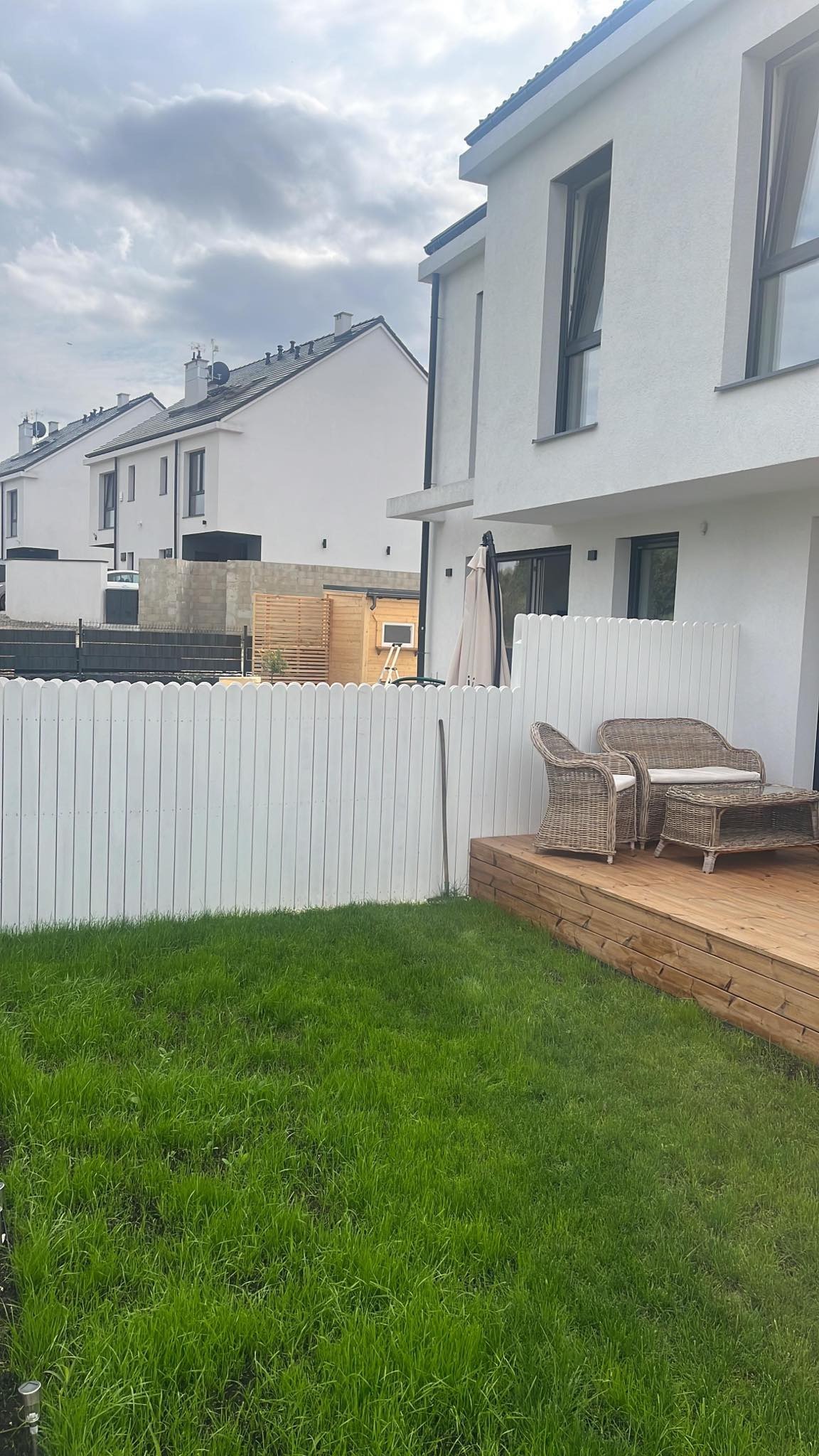 Drewniany taras z rattanowym zestawem mebli ogrodowych i białym płotem na tle nowoczesnych domów w zabudowie szeregowej, trawa po koszeniu.