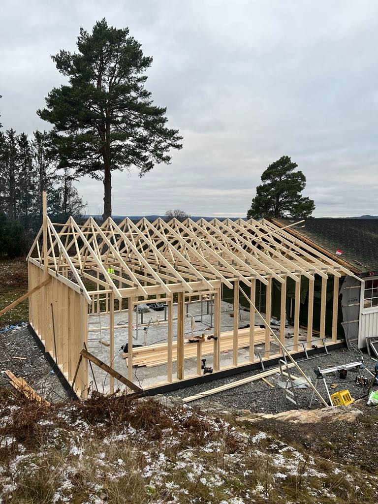 Konstrukcja szkieletowa budynku w trakcie budowy, widoczne drewniane ramy ścian i dachu na betonowej płycie fundamentowej, teren wokół częściowo pokryty śniegiem, pochmurne niebo w tle.