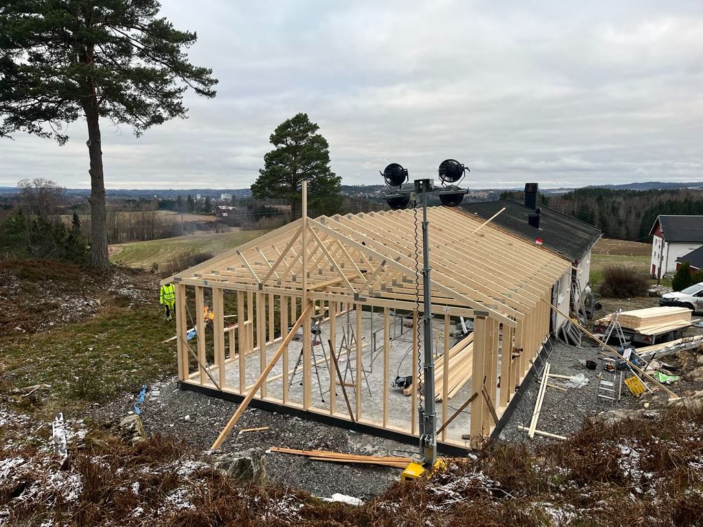 Budowa domu szkieletowego w trakcie: widoczna drewniana konstrukcja, zarys ścian i dachu, teren budowy z elementami krajobrazu i sprzętem budowlanym, pochmurne niebo w tle.