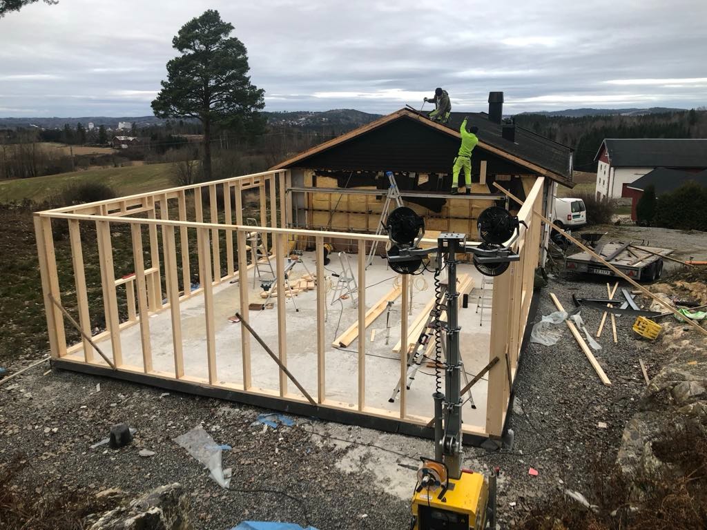 Wznoszenie szkieletu domu jednorodzinnego, widoczna drewniana konstrukcja ścian, ocieplenie dachu, pracownicy na dachu, teren budowy z narzędziami i oświetleniem.