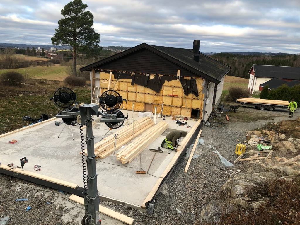 Budowa domu: Widok z góry na plac budowy z fundamentami, izolacją ścian i stosem drewna, z pracownikiem w zielonym kombinezonie przycinającym drewno, w tle krajobraz z drzewami i zabudowaniami.