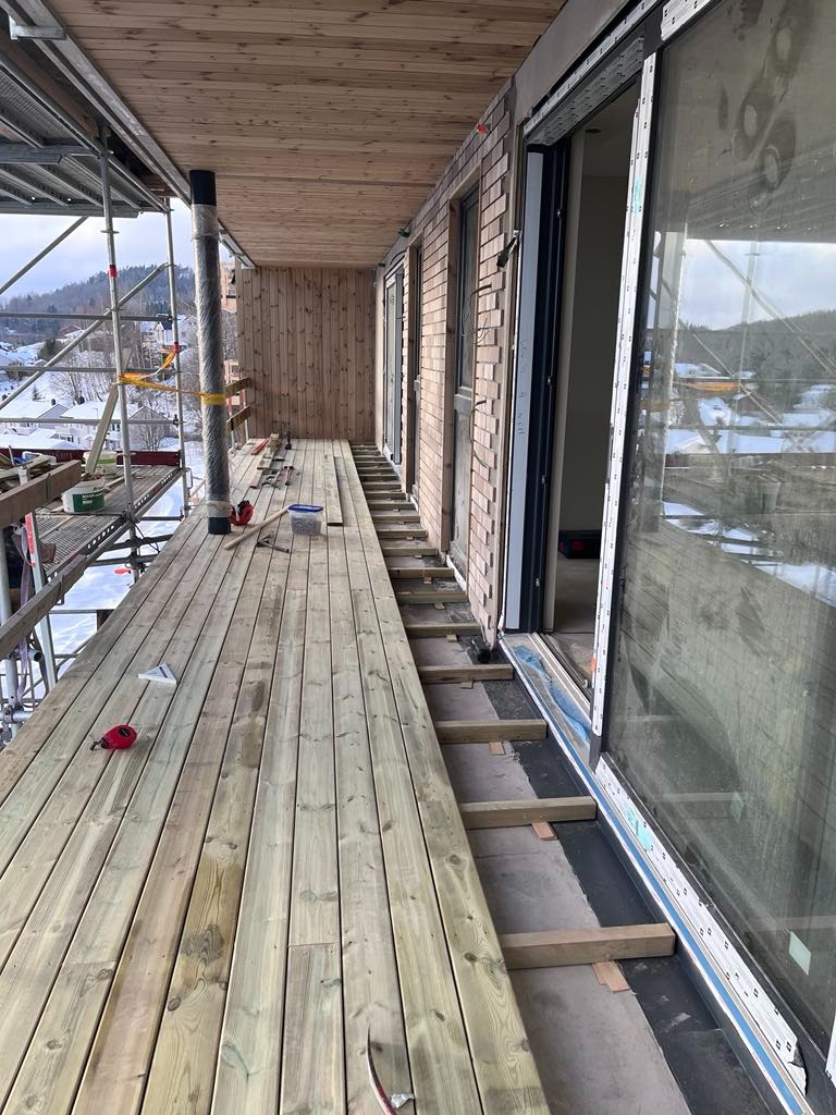 Montaż drewnianego tarasu przy domu szkieletowym, widoczne deski podłogowe, rusztowanie i narzędzia pracy.