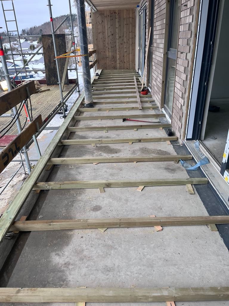 W trakcie budowy tarasu: widoczne drewniane legary ułożone na betonowej wylewce, w tle rusztowania i budynek w trakcie wykończenia elewacji drewnianej.