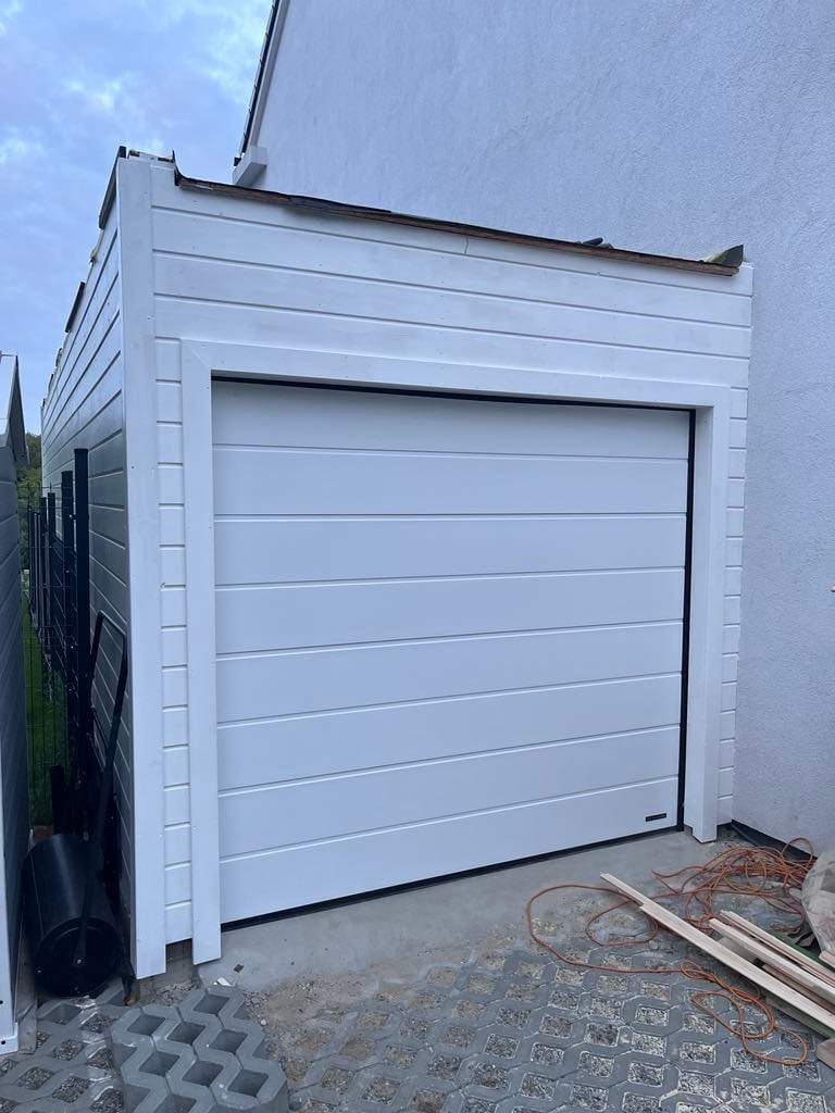 Nowoczesny, biały garaż z poziomymi panelami, przylegający do budynku, z widocznymi elementami budowlanymi i narzędziami na betonowej posadzce.