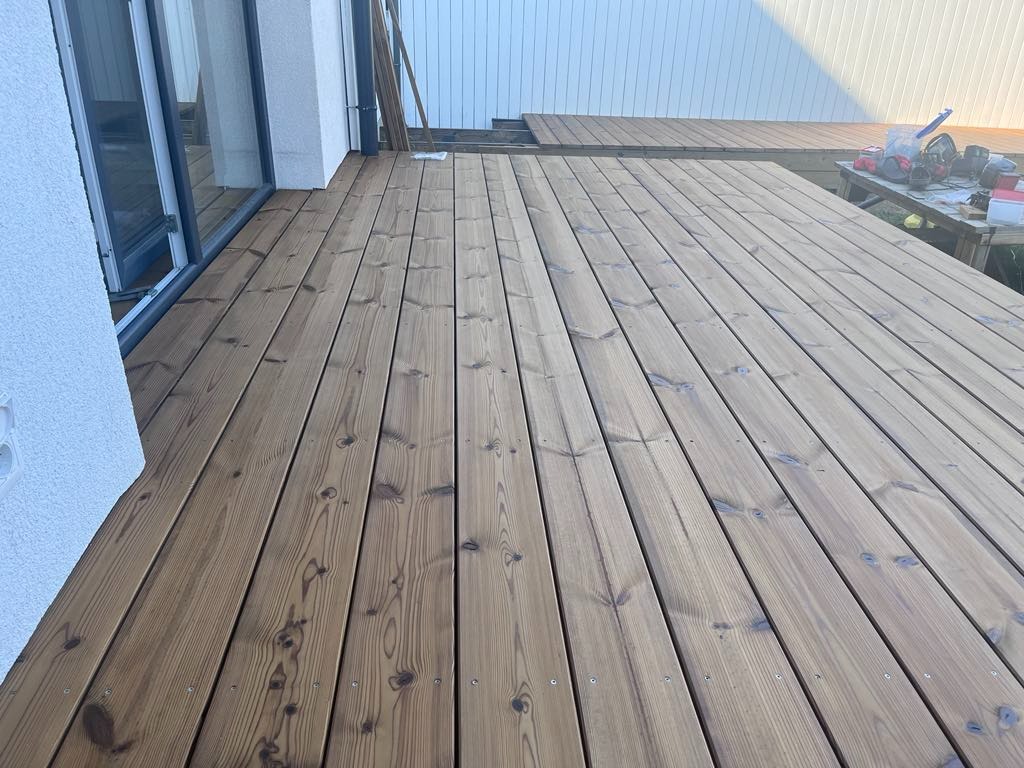 Świeżo zaimpregnowany drewniany taras przylegający do białej ściany budynku, z widocznym fragmentem elewacji i narzędziami na stole w tle.