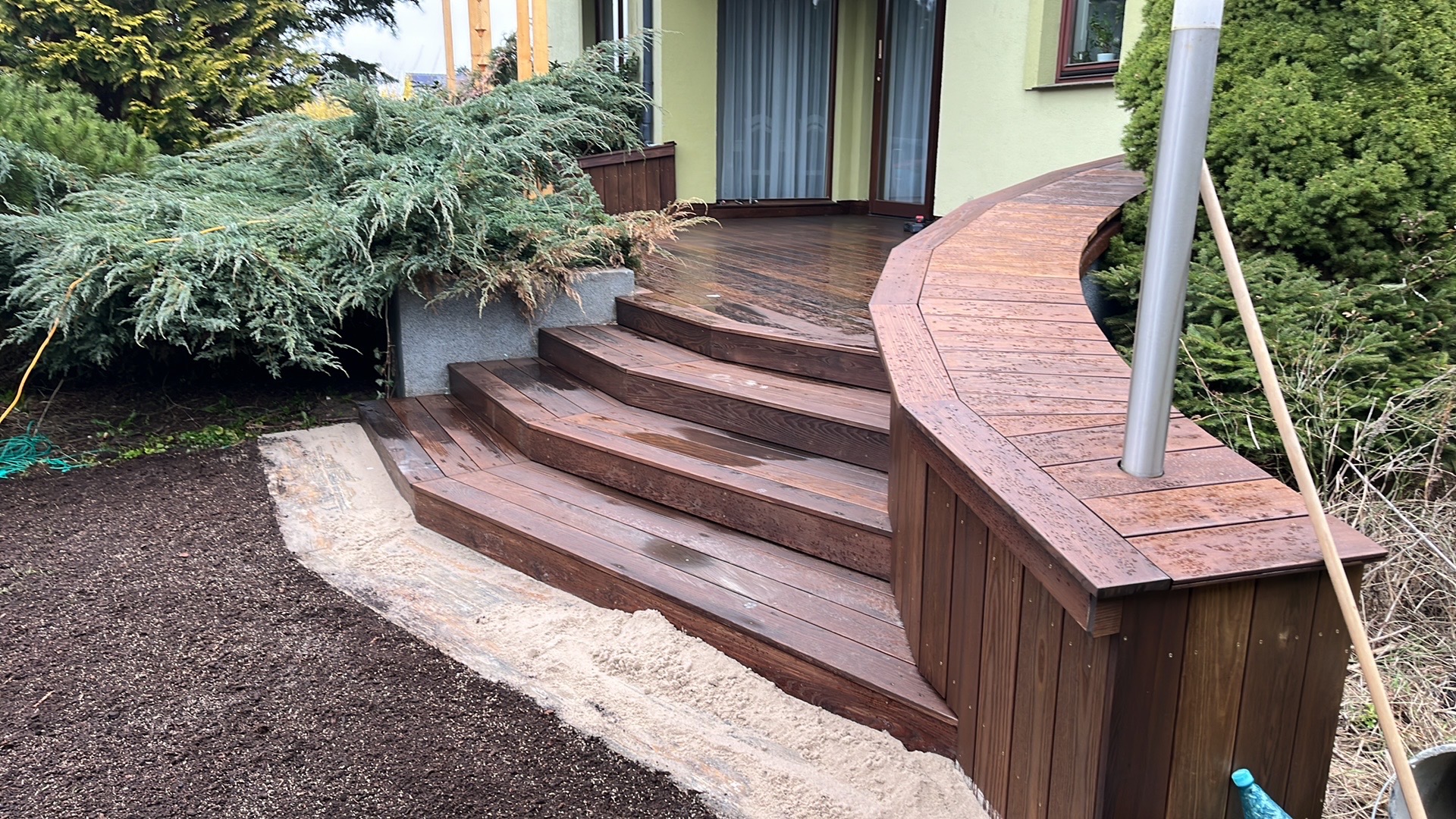 Schody i fragment tarasu z ciemnego drewna, z widocznymi kroplami deszczu, prowadzące do domu z zieloną elewacją, obok świeżo przygotowany teren pod nasadzenia.