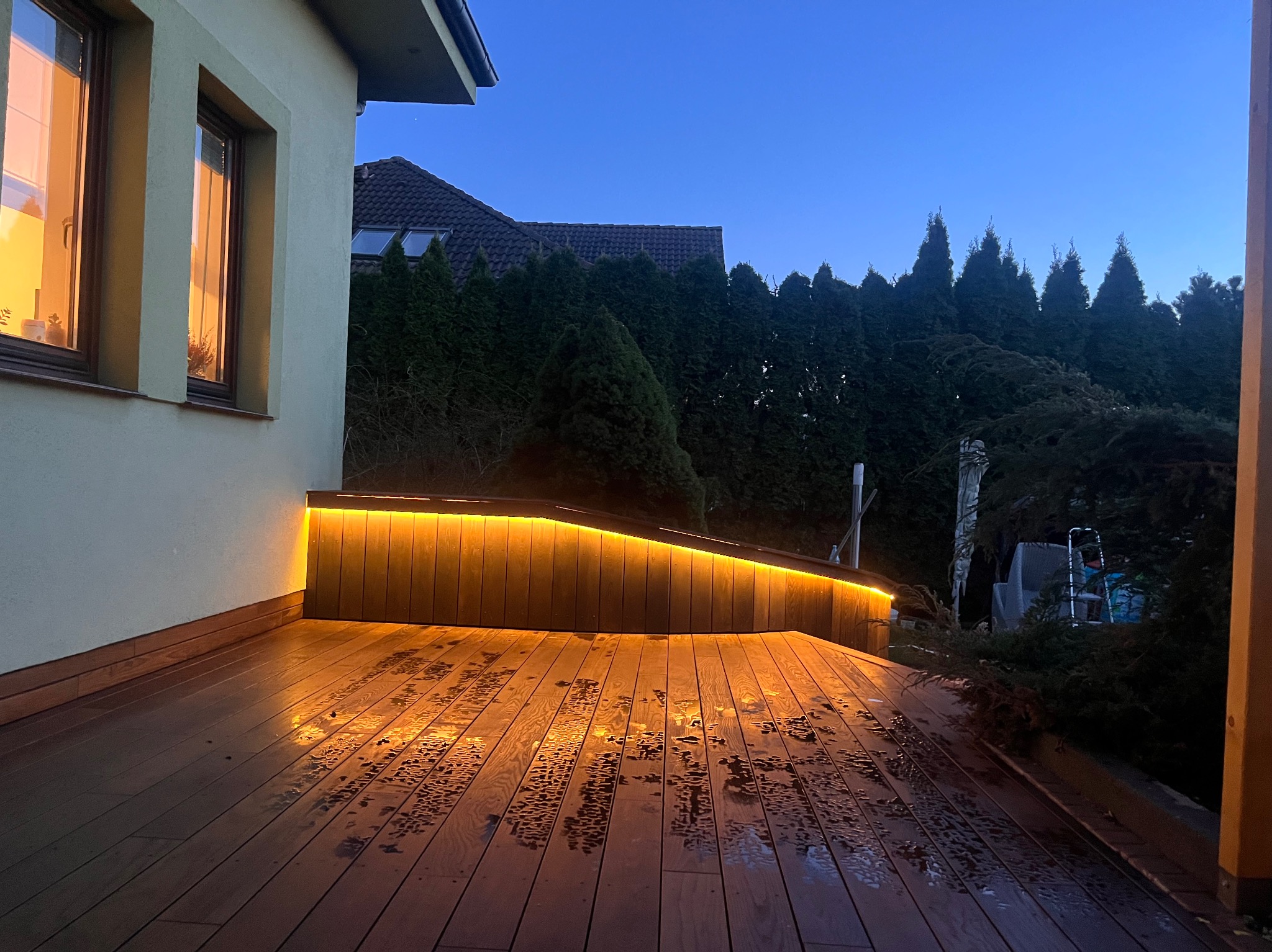 Oświetlony taras drewniany po deszczu, widok z poziomu podłogi, z podświetlaną listwą LED wzdłuż krawędzi, na tle ciemnego nieba i zielonych krzewów.