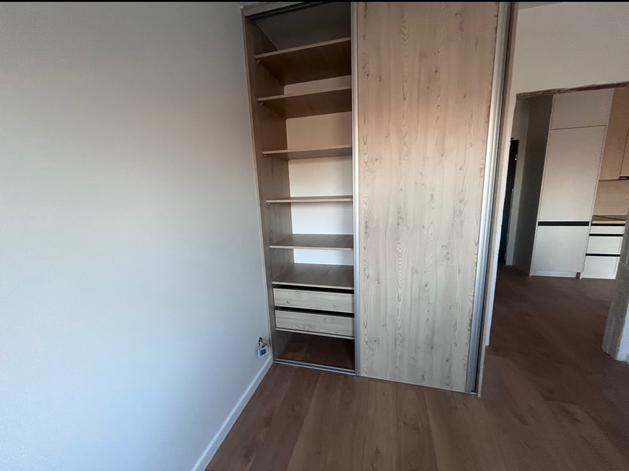 Wnętrze szafy przesuwnej z półkami i szufladami, widok od frontu, drzwi przesuwne częściowo zasłonięte, podłoga drewniana, fragment ściany w tle.