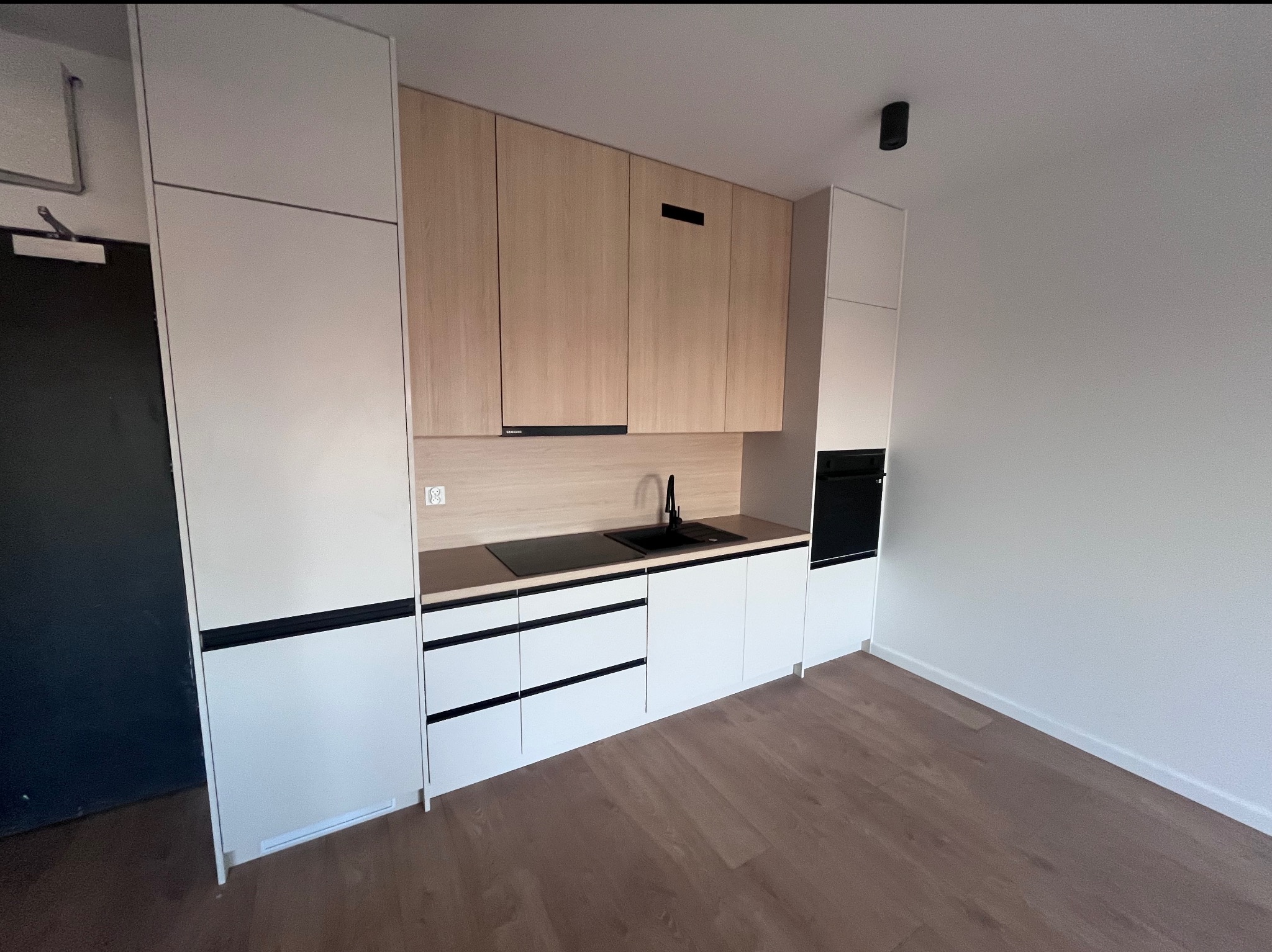 Nowoczesna zabudowa kuchenna z frontami w kolorze jasnego drewna i białymi szafkami, czarny zlew i płyta grzewcza, drewniana podłoga, minimalistyczny design.