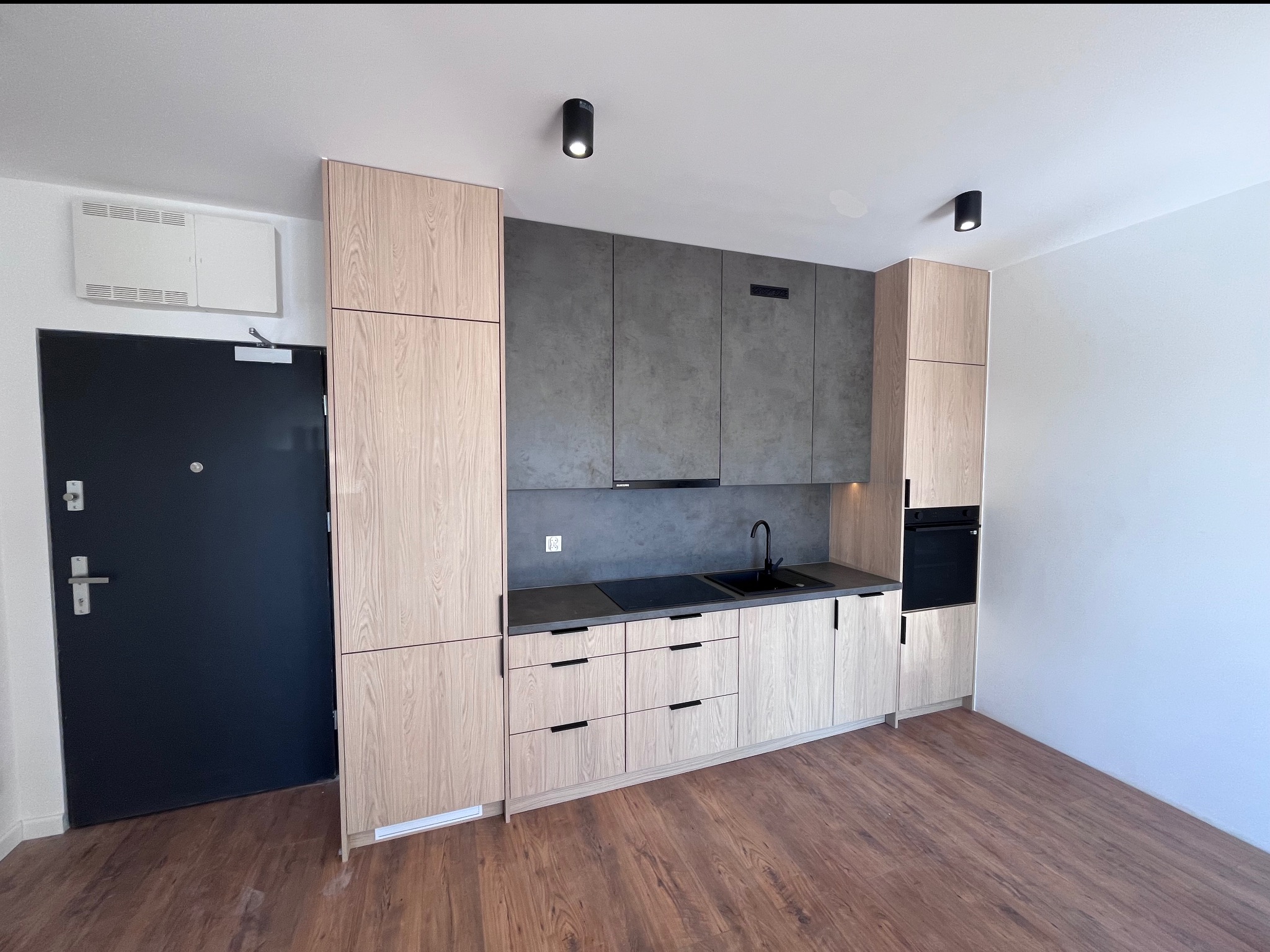 Nowoczesna kuchnia z drewnianymi szafkami i szarym blatem, czarna armatura i oświetlenie punktowe, widok na drzwi wejściowe.