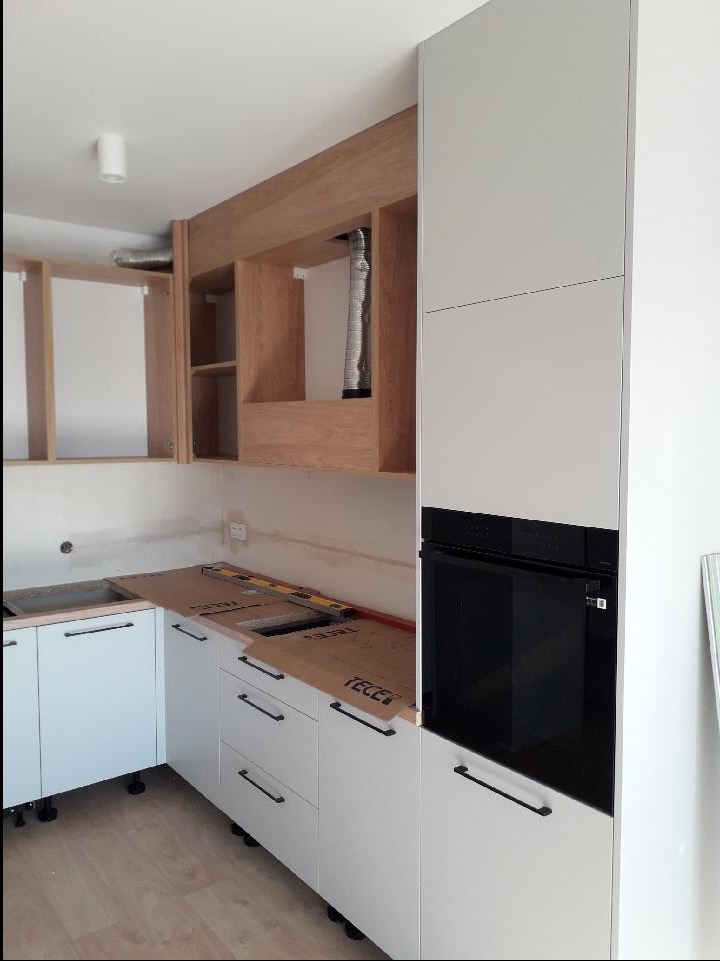 Montaż kuchni na wymiar: białe szafki z czarnymi uchwytami, zabudowana piekarnik, drewniane szafki wiszące bez frontów, widoczne elementy instalacji wentylacyjnej, blat zabezpieczony kartonem...