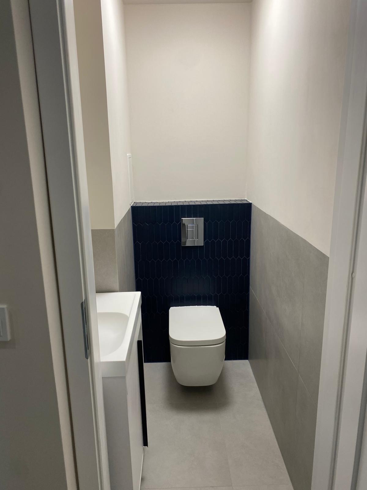Nowoczesna toaleta po remoncie: biała ceramika, granatowe płytki o unikalnym kształcie i szare ściany. Minimalistyczny design, widok zza uchylonych drzwi.