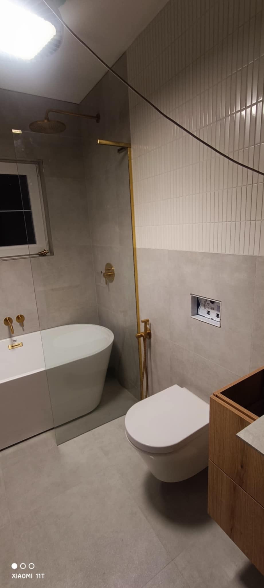 Wykończona łazienka z wanną wolnostojącą, toaletą podwieszaną i złotą armaturą. Na ścianach szare płytki z białym pasem. Widoczne okno i fragment szafki pod umywalkę.