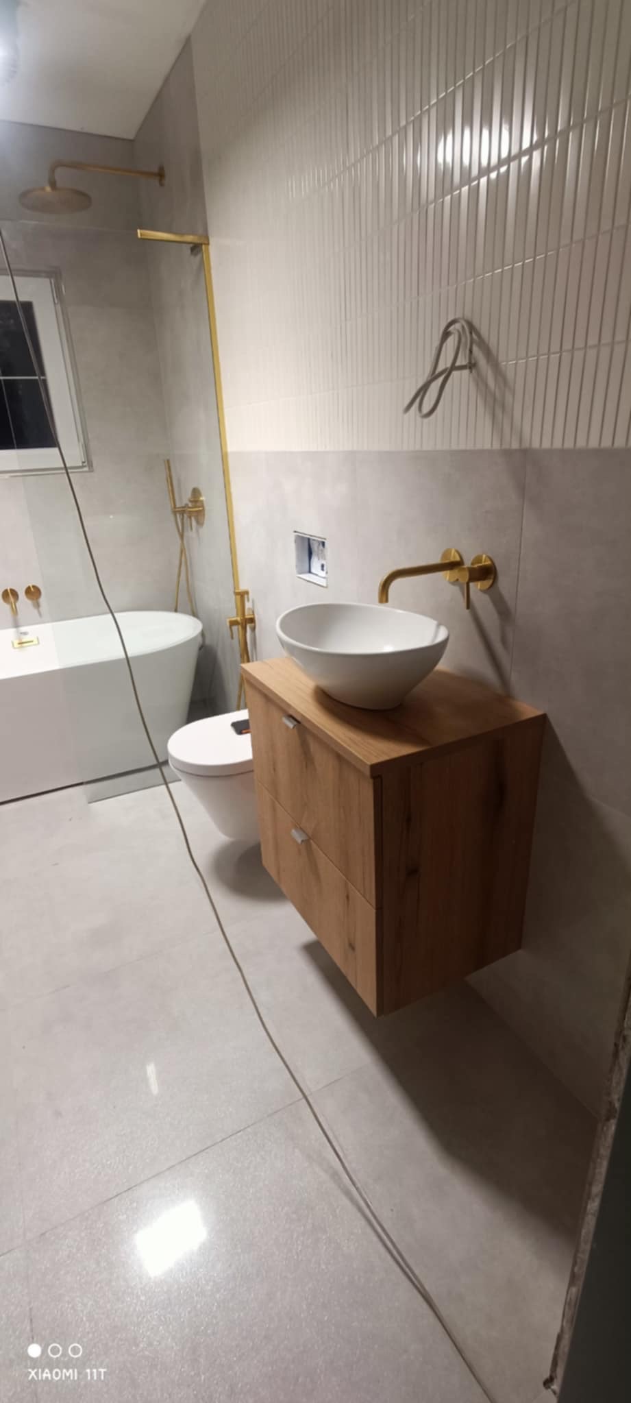 Nowoczesna łazienka z białą umywalką na drewnianej szafce, złotą armaturą i wanną. Ściany wyłożone płytkami w odcieniach szarości. Widoczne elementy instalacji.