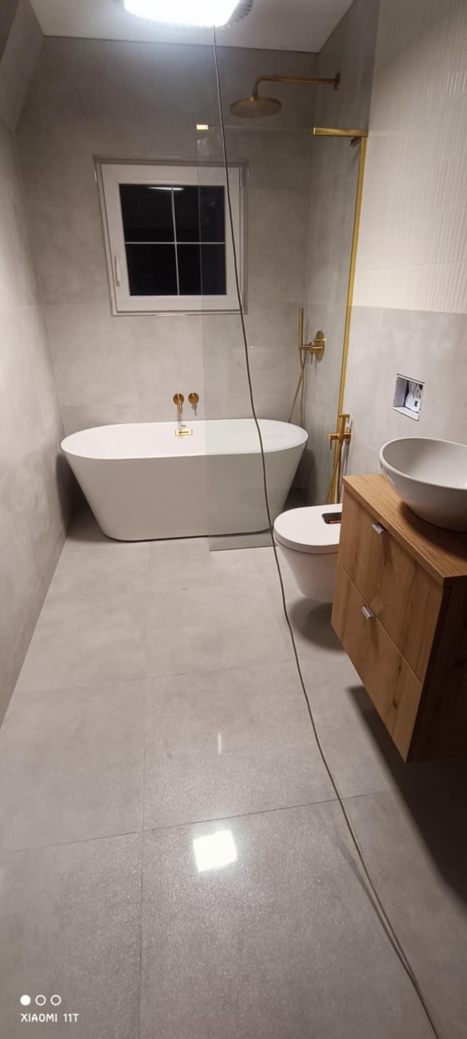 Nowoczesna łazienka w szarościach z wolnostojącą wanną, złotą armaturą i drewnianą szafką pod umywalką. Okno z białymi ramami doświetla wnętrze.