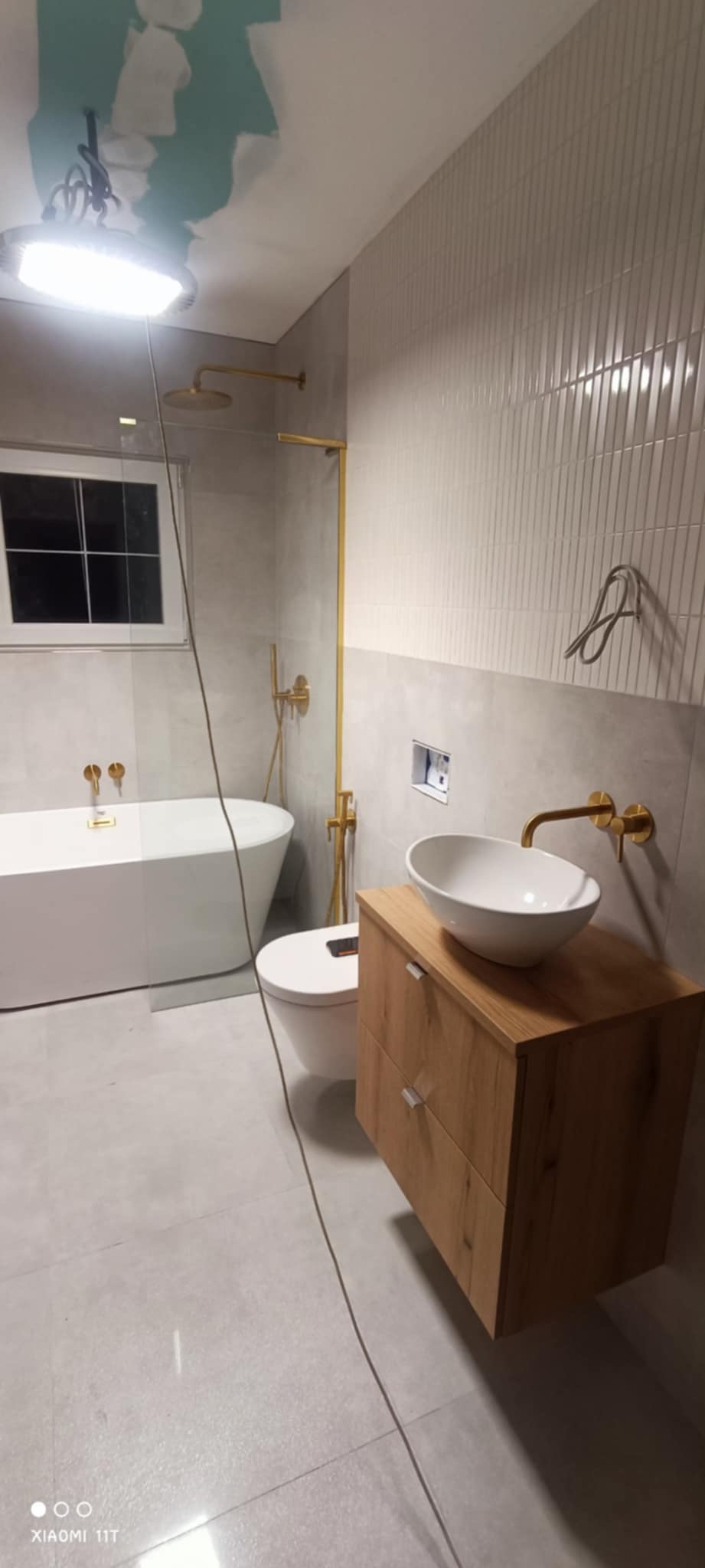 Nowoczesna łazienka z wolnostojącą wanną, złotą armaturą i drewnianą szafką pod umywalką. Ściany wyłożone białymi, wąskimi płytkami.