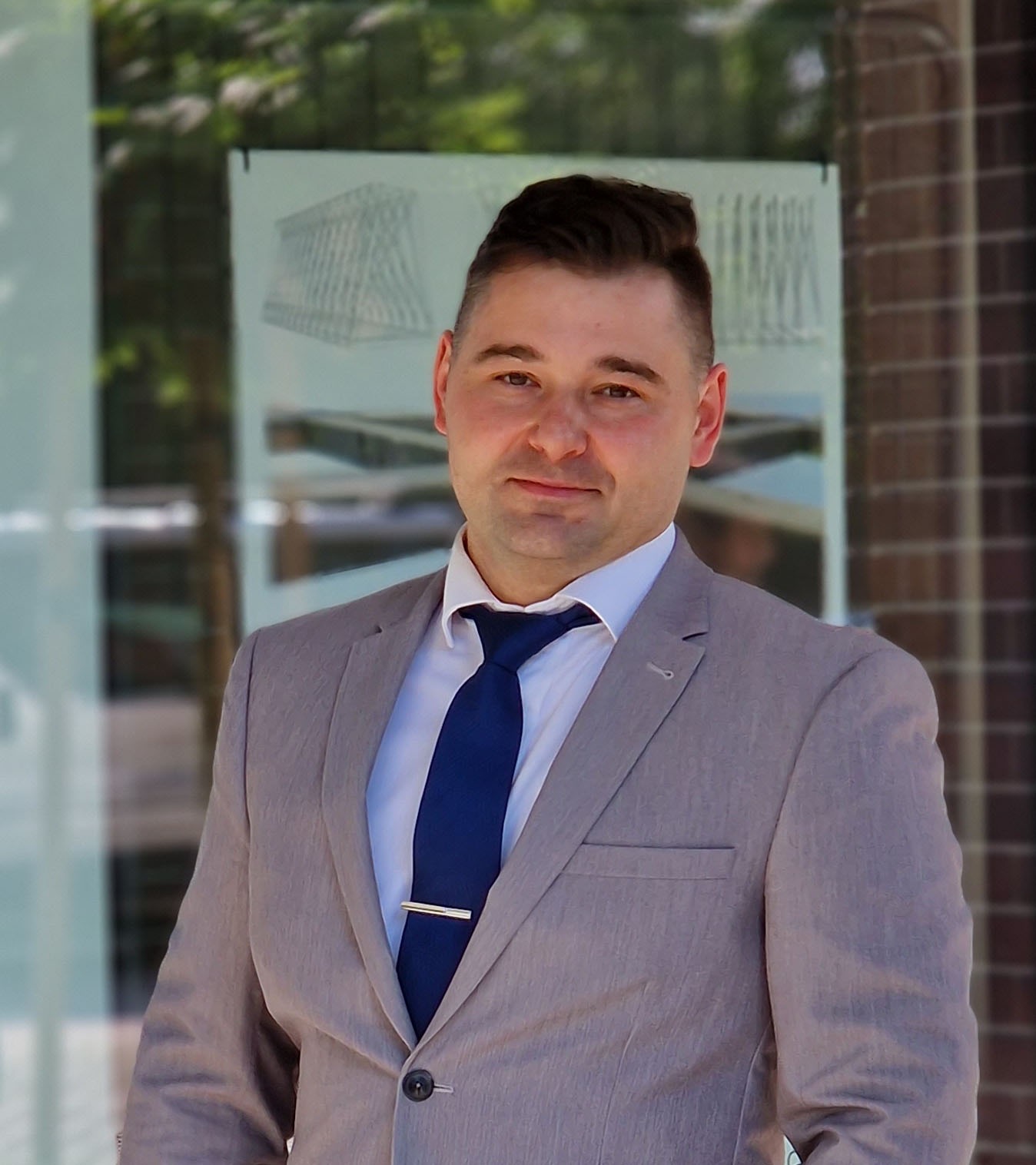 Mężczyzna w szarym garniturze i niebieskim krawacie, na tle przeszklonej ściany z widocznym rysunkiem architektonicznym budynku.