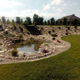 Mariusz Radwański Usługi Ogrodnicze - Ogród w Słupsku z oczkiem wodnym otoczonym kamieniami, kaskada wodna i schody prowadzące na wyższy poziom ogrodu, trawnik.