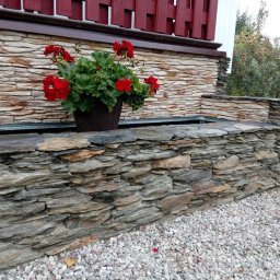 Mariusz Radwański Usługi Ogrodnicze - Kamienny murek z kwiatami w donicy, wykonany z nieregularnych płatów kamienia, otoczony białym żwirem.