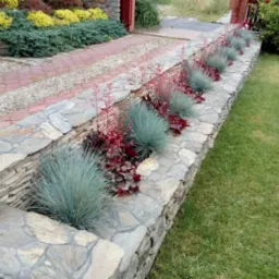 Kamienna rabata z ozdobnymi trawami i czerwonymi kwiatami, oddzielająca trawnik od ścieżki z kostki i żwiru.