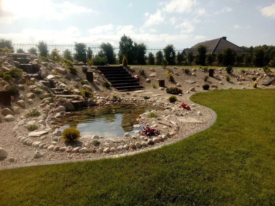 Ogród w Słupsku z oczkiem wodnym otoczonym kamieniami, kaskada wodna i schody prowadzące na wyższy poziom ogrodu, trawnik.