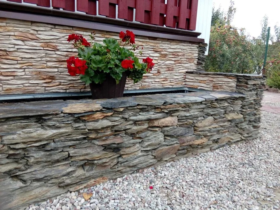 Kamienny murek z kwiatami w donicy, wykonany z nieregularnych płatów kamienia, otoczony białym żwirem.