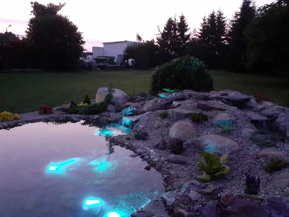 Oświetlony, kaskadowy zbiornik wodny z kamieniami i roślinami, w tle trawnik i budynek gospodarczy.