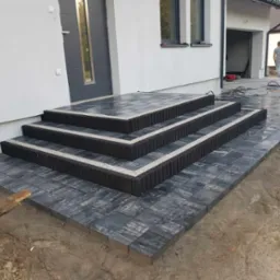 Nowoczesne schody wejściowe z ciemnej kostki brukowej z jasnymi akcentami, prowadzące do domu z szarą stolarką okienną i drzwiami, widoczne prace wykończeniowe.