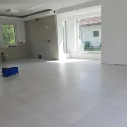 Jasne, duże płytki podłogowe w nowym, przestronnym pomieszczeniu z dwoma oknami, ściany częściowo w stanie surowym, niebieski w&oacute;zek z narzędziami w rogu, widoczne elementy instalacji elektrycznej.