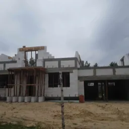 Niedokończony dom murowany z betonu komórkowego, widoczne drewniane podpory i szalunki, cegły na ziemi, w tle drzewa.