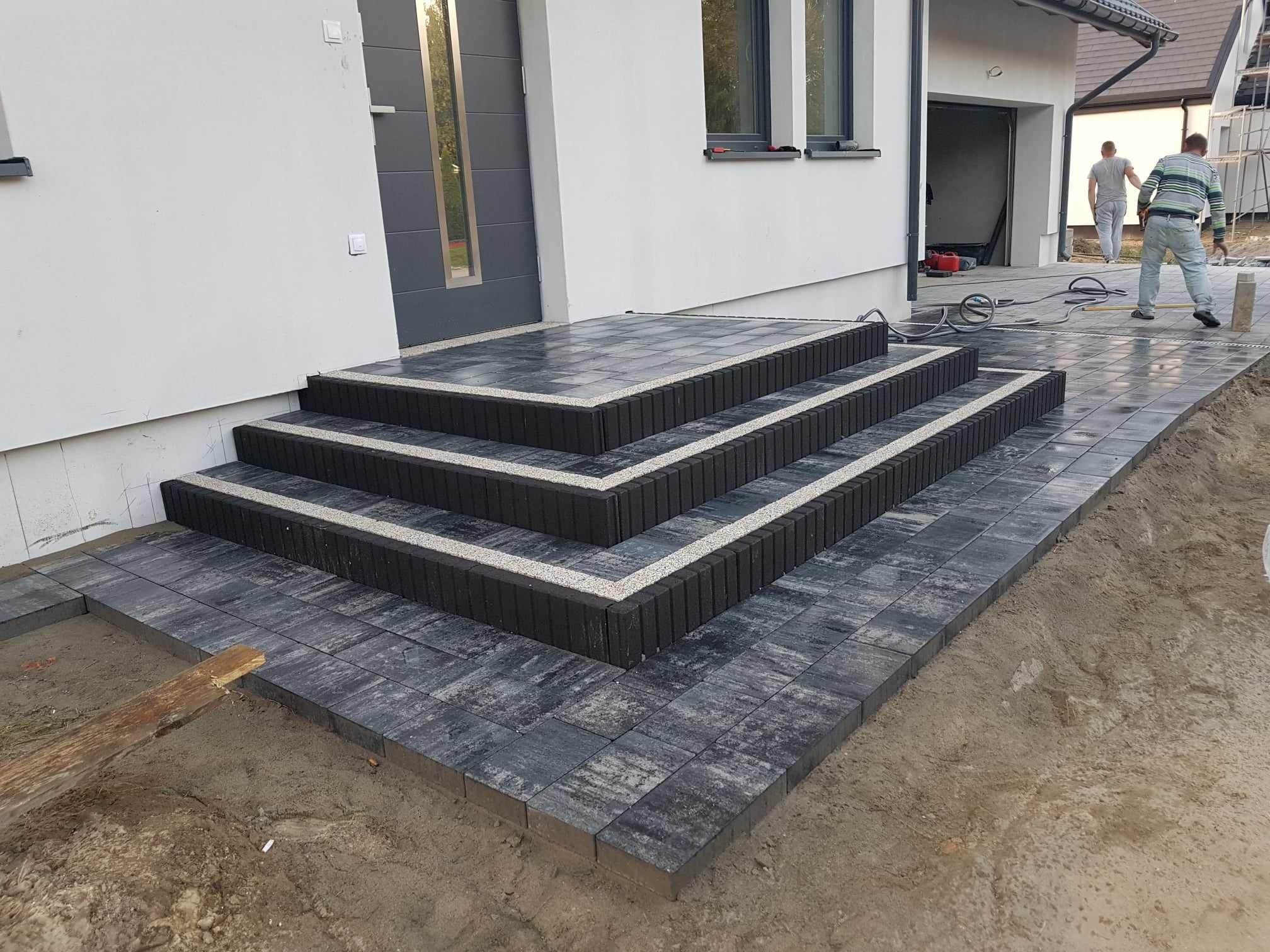 Nowoczesne schody wejściowe z ciemnej kostki brukowej z jasnymi akcentami, prowadzące do domu z szarą stolarką okienną i drzwiami, widoczne prace wykończeniowe.