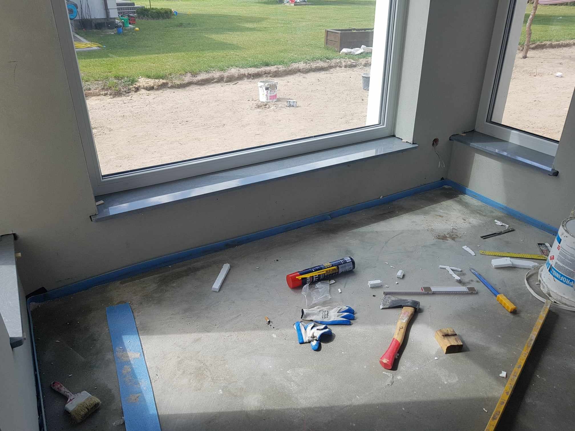 Wnętrze w trakcie prac wykończeniowych: osadzone parapety okienne, zabezpieczona taśmą podłoga, widoczne narzędzia glazurnicze i budowlane, w tle trawnik za oknem.