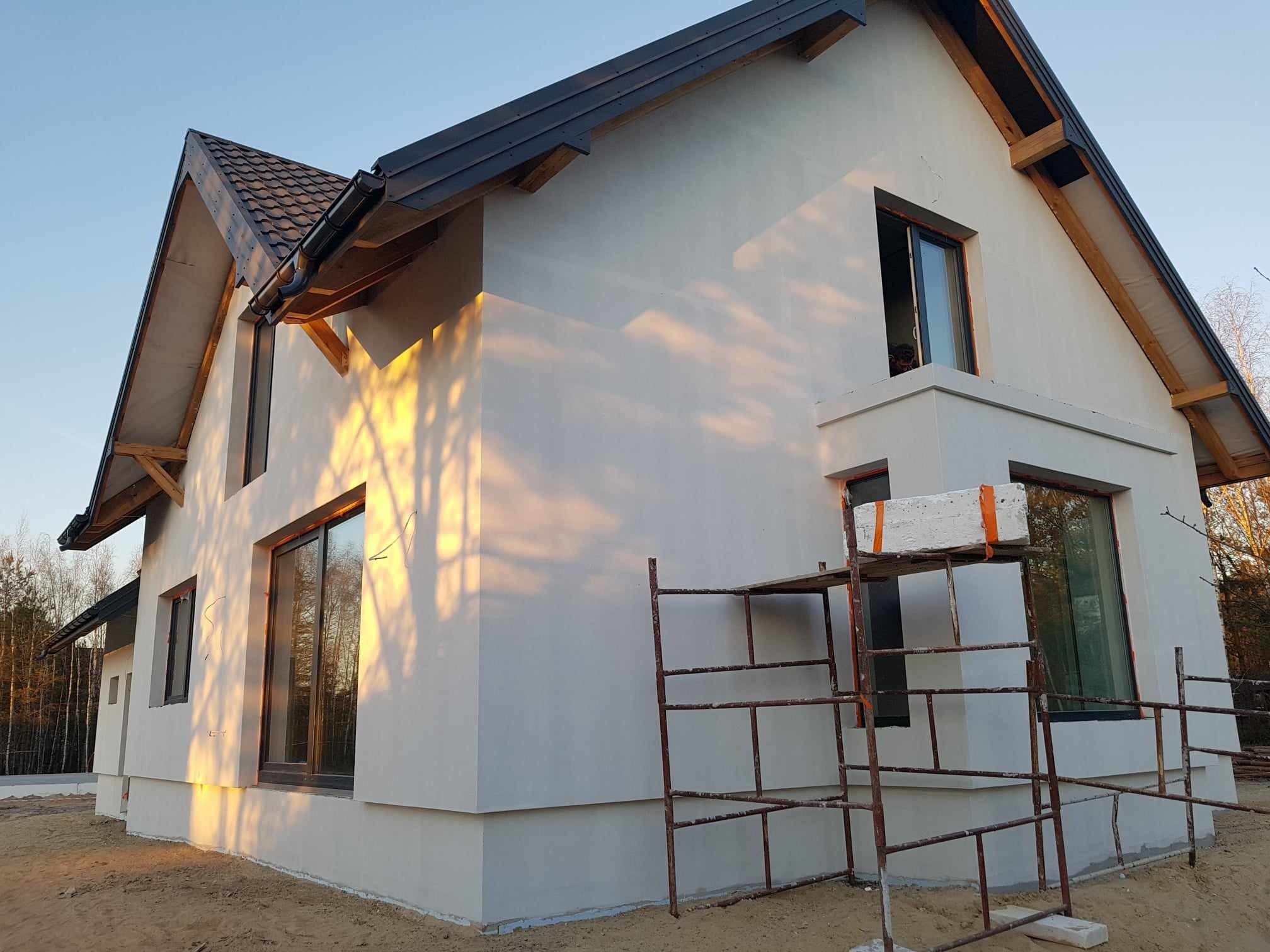 Surowy dom jednorodzinny w trakcie wykończenia, z widocznym rusztowaniem i świeżo nałożonym tynkiem zewnętrznym, oświetlony popołudniowym słońcem rzucającym cienie drzew na ścianę.