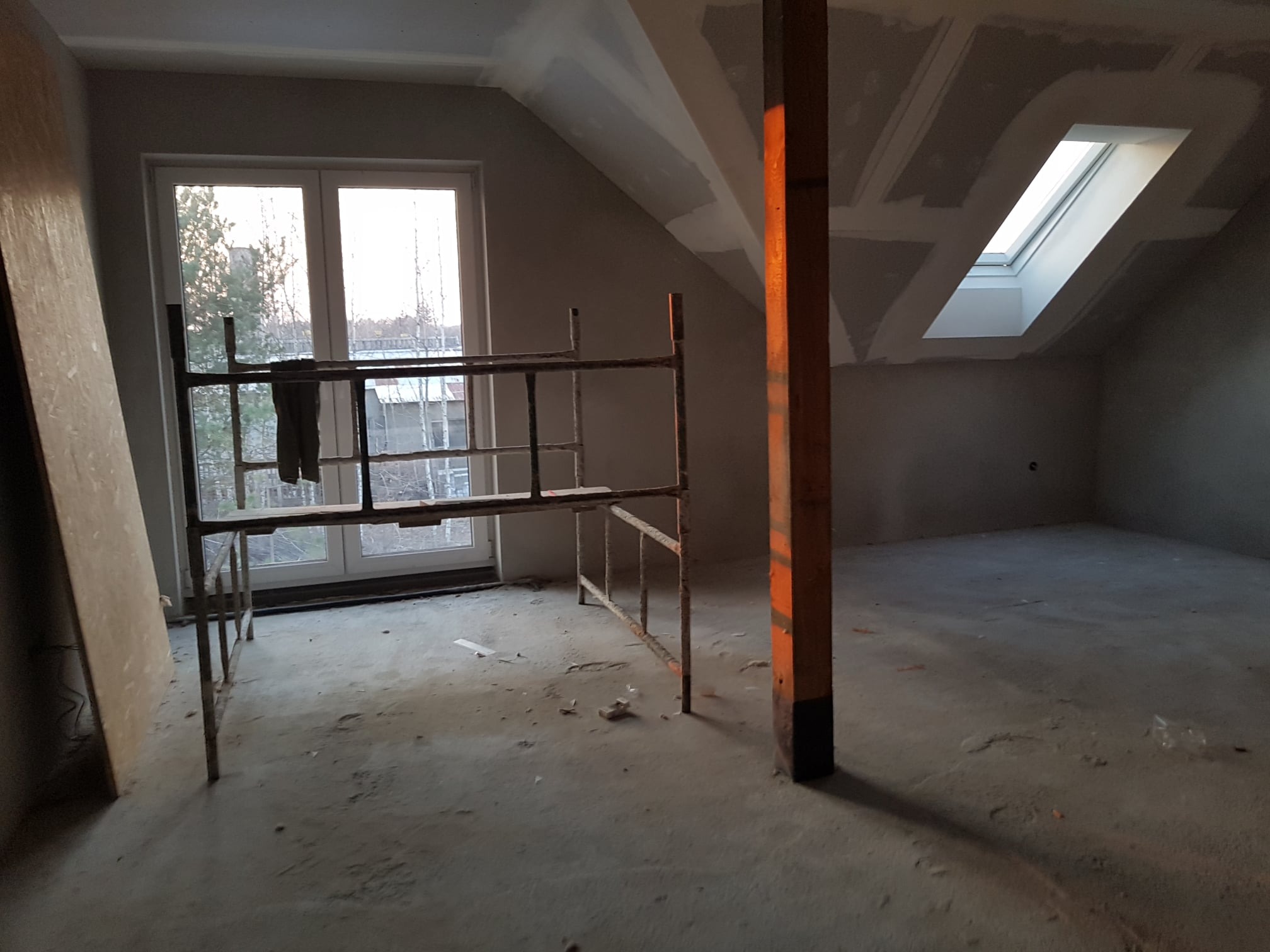 Poddasze w trakcie wykańczania: widoczne rusztowanie, okno dachowe, okno w ścianie szczytowej, częściowo wykończone ściany płytami kartonowo-gipsowymi ze śladami szpachlowania, drewniany słup...