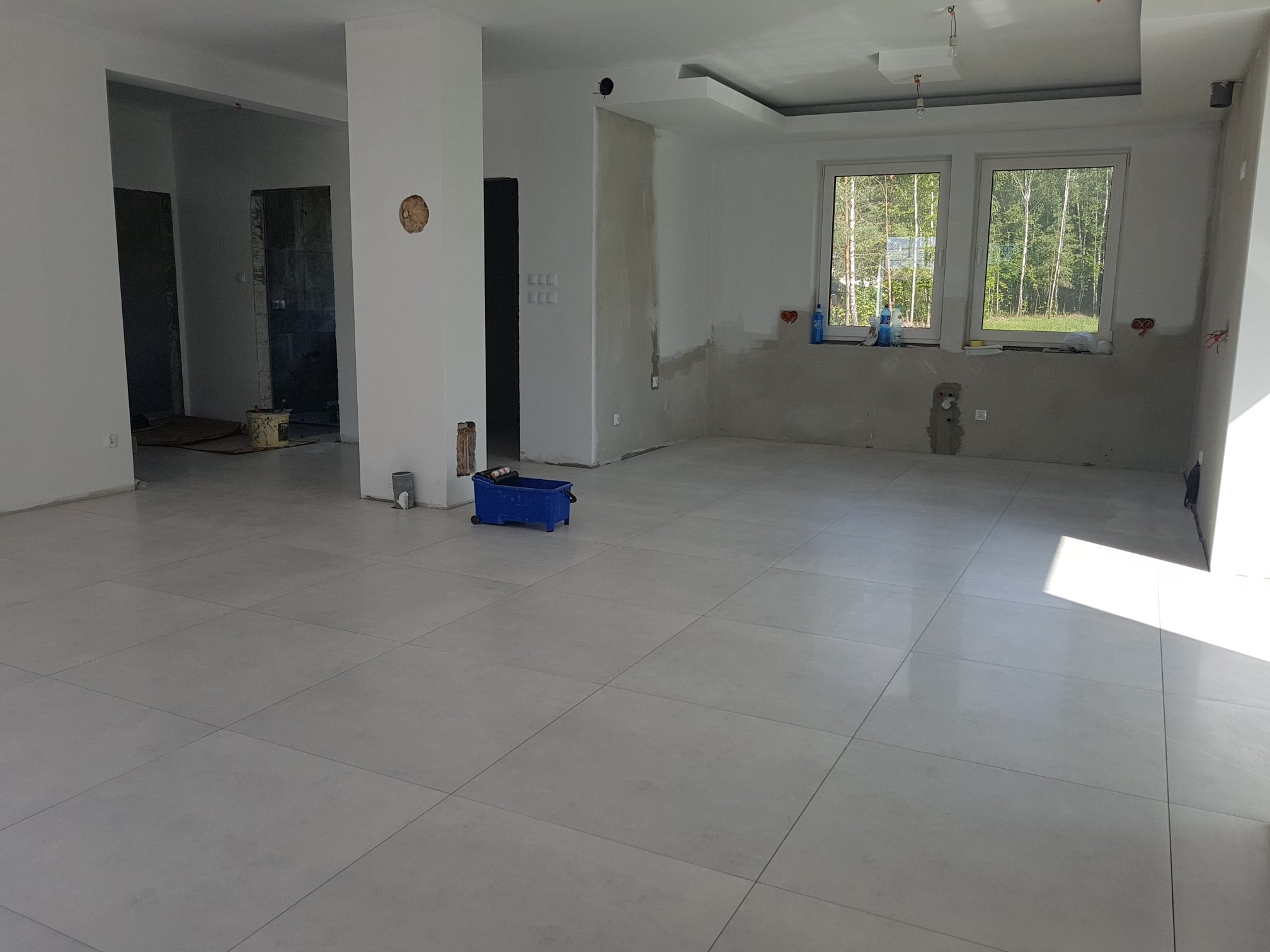 Wnętrze w trakcie wykańczania: jasne płytki podłogowe, ściany częściowo wyszpachlowane, widoczne puszki elektryczne, okna z widokiem na zieleń, niebieski wózek budowlany.