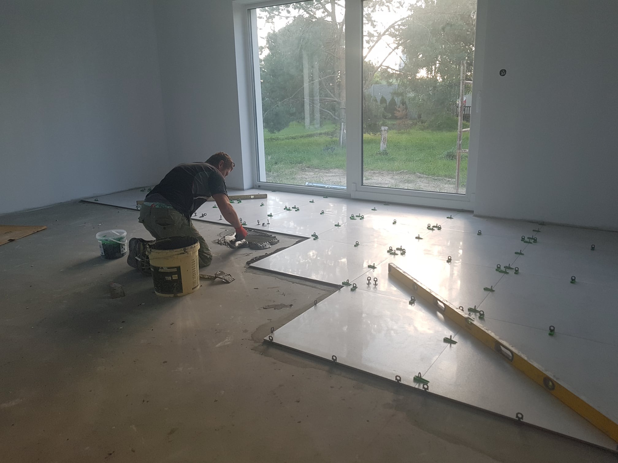 Układanie błyszczących, jasnych płytek podłogowych w dużym pokoju z widokiem na ogród przez duże okno; widoczne narzędzia glazurnicze, poziomica i system poziomowania płytek.