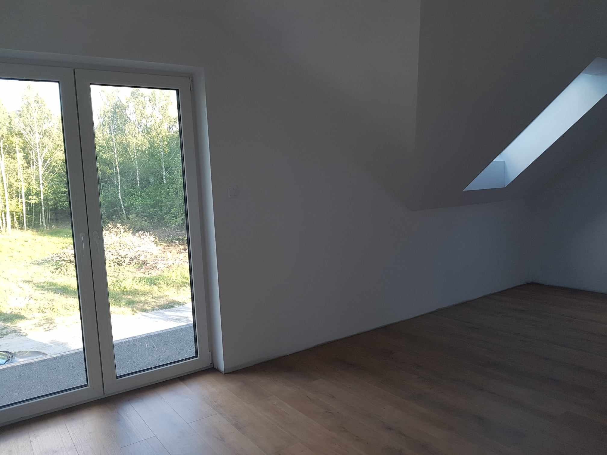 Poddasze z oknem dachowym i dużym oknem balkonowym z widokiem na drzewa, świeżo wykończone, z drewnianą podłogą.