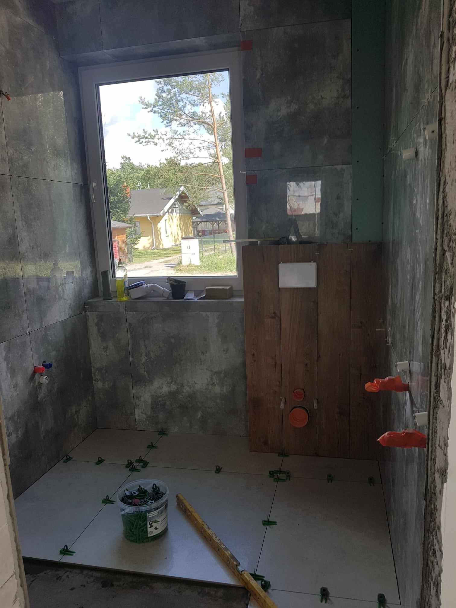 Wnętrze łazienki w trakcie układania szarych płytek ceramicznych w Białej Podlaskiej, widoczne krzyżyki dystansowe, instalacja podtynkowa WC oraz okno z widokiem na dom.