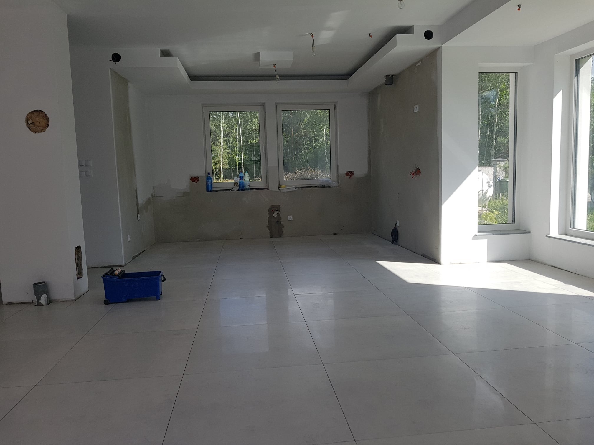 Wnętrze w stanie deweloperskim z ułożoną jasną, kwadratową podłogą z płytek, widoczne ślady po pracach wykończeniowych na ścianach, niebieski pojemnik na kółkach i okna z widokiem na zieleń.