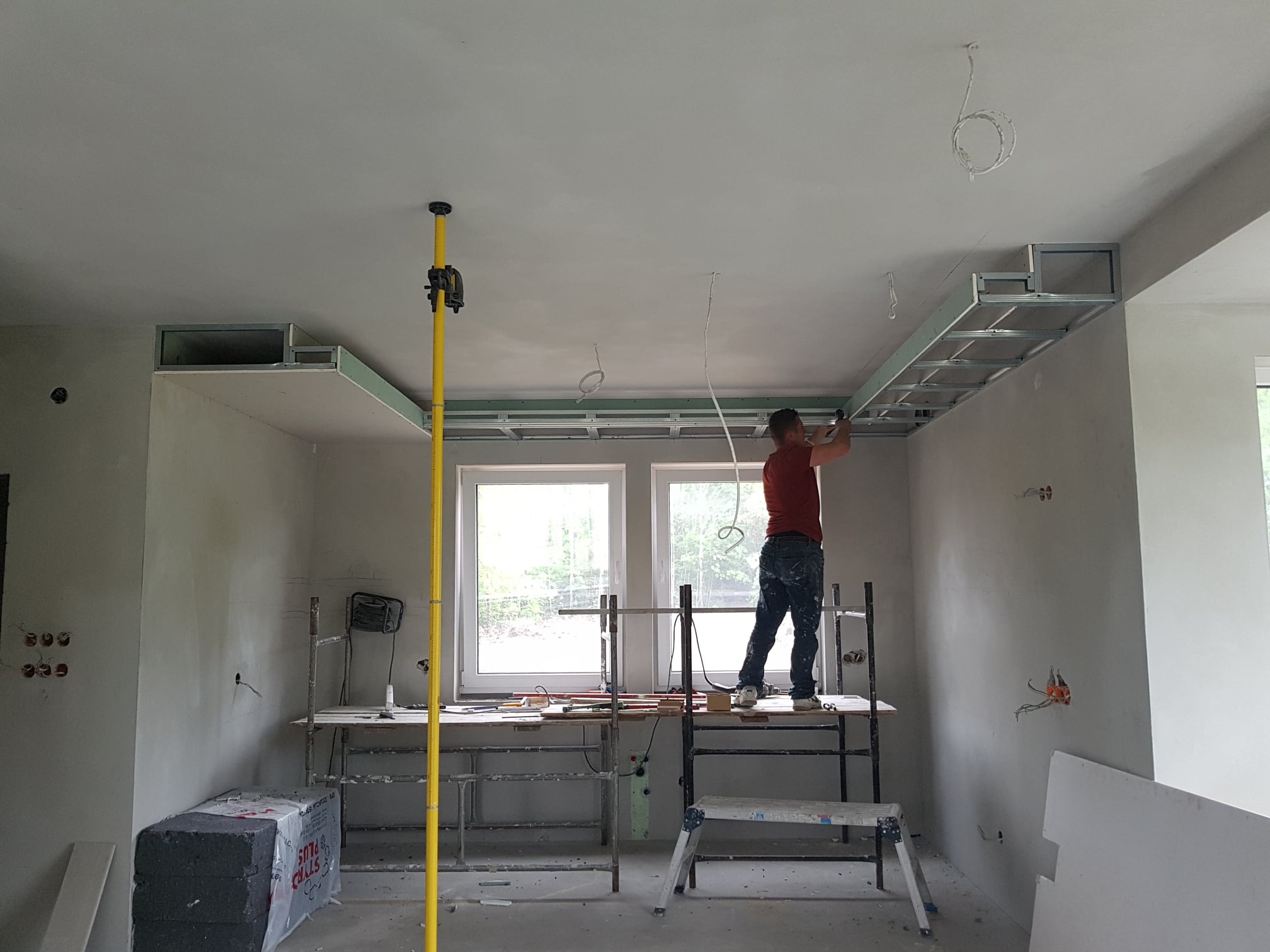 Pracownik na rusztowaniu montuje metalowy stelaż sufitu podwieszanego z płyt gipsowo-kartonowych w pomieszczeniu z oknami, widoczne przewody elektryczne i laserowy niwelator pionowy.
