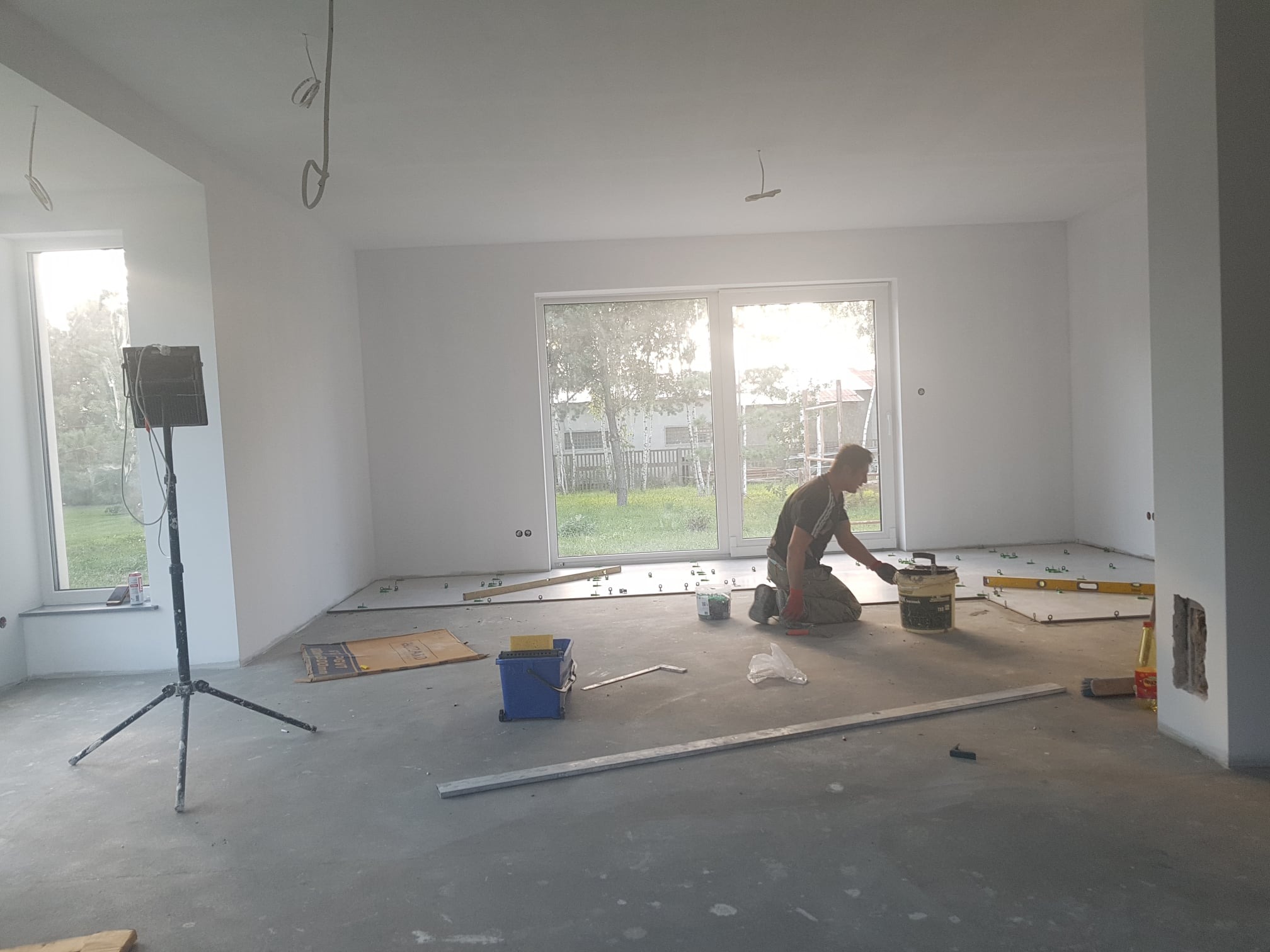 Glazurnik układa płytki podłogowe w jasnym, przestronnym pomieszczeniu z dużym oknem, widoczne narzędzia i poziomice.