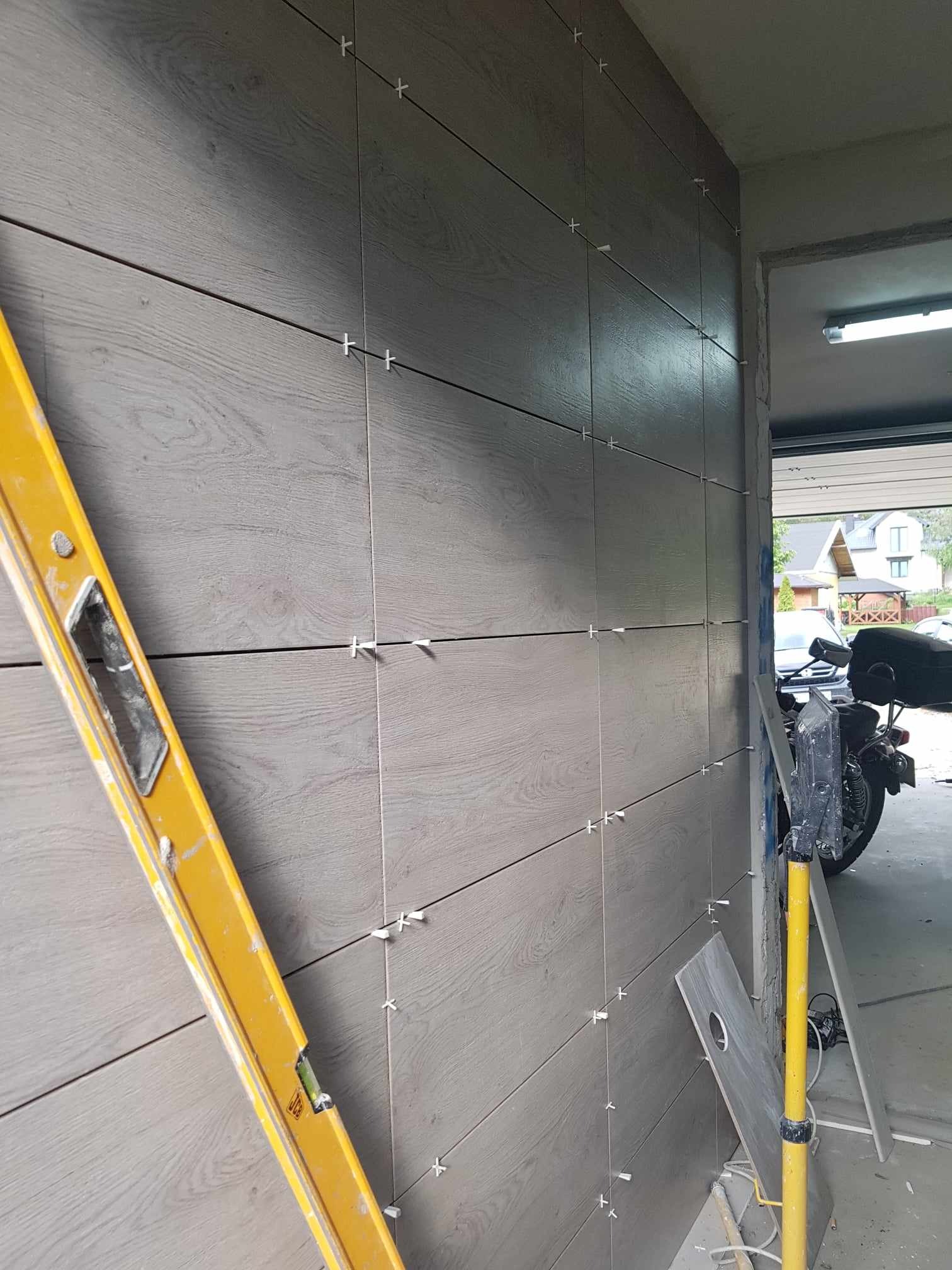 Ściana wyłożona płytkami imitującymi drewno w trakcie montażu, widoczne krzyżyki dystansowe, poziomica i sprzęt glazurniczy, w tle garaż z motocyklem.